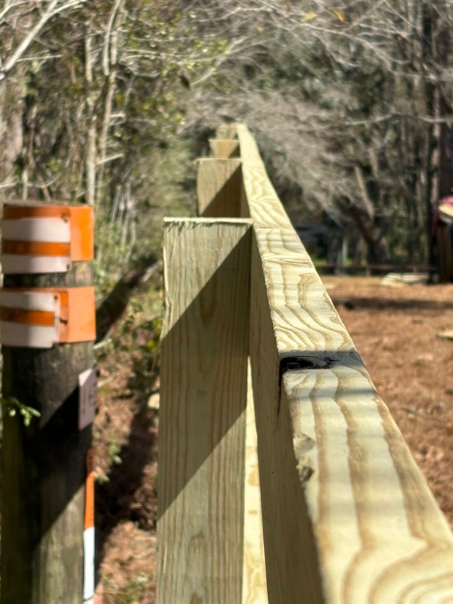 A perspective view along the top edge of a new wooden fence, with a striped orange and white post in the foreground.