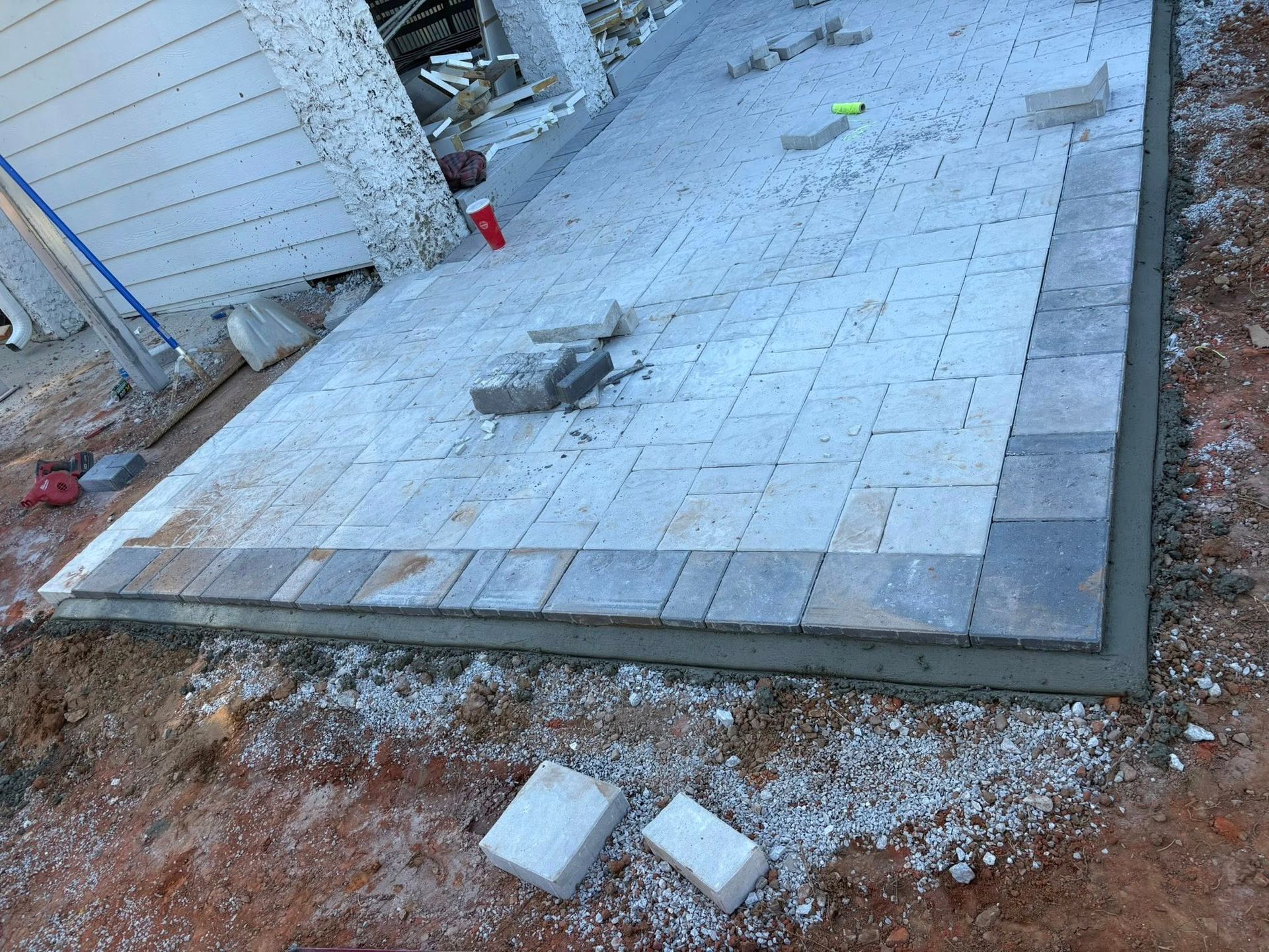 A partially finished rectangular stone paver patio sits on a bed of gravel next to the exterior wall of a house.