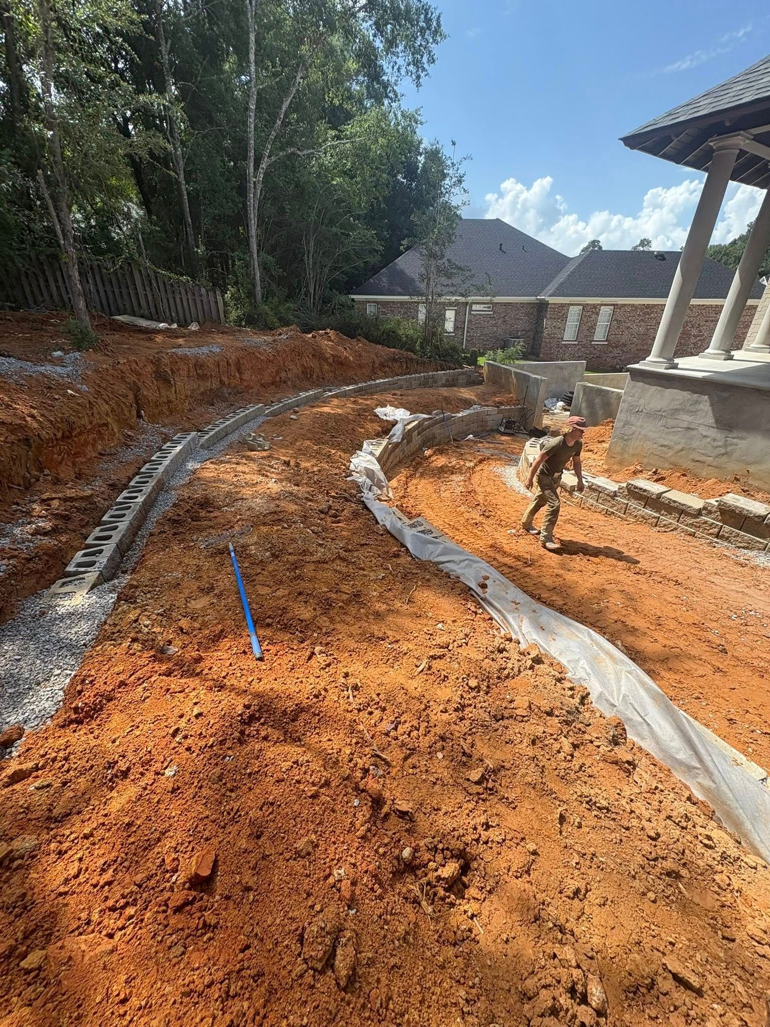 A person walks near a retaining wall under construction in a large, red-dirt yard next to a house.