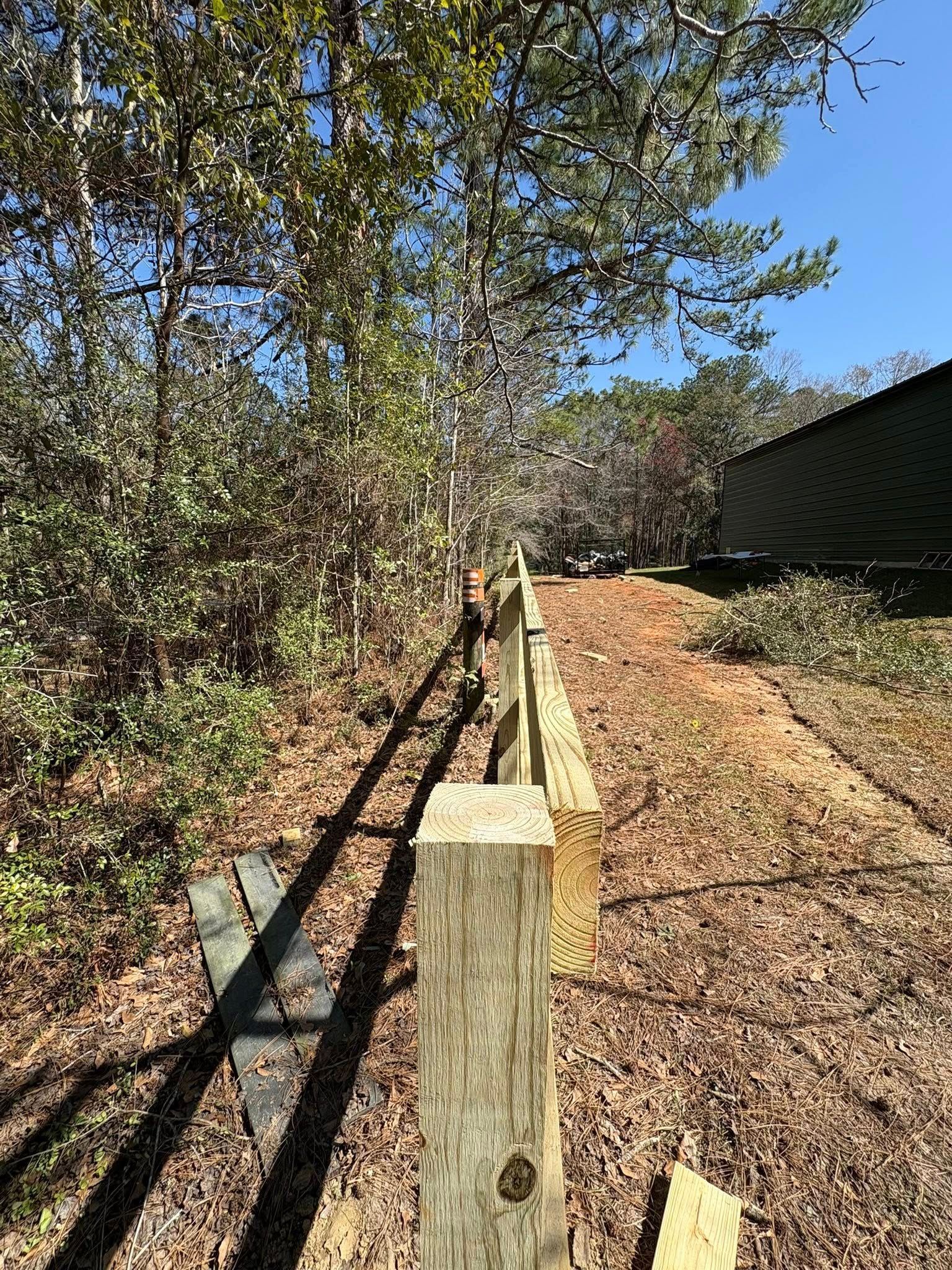 A wooden post-and-rail fence under construction along a wooded edge on a sunny day.