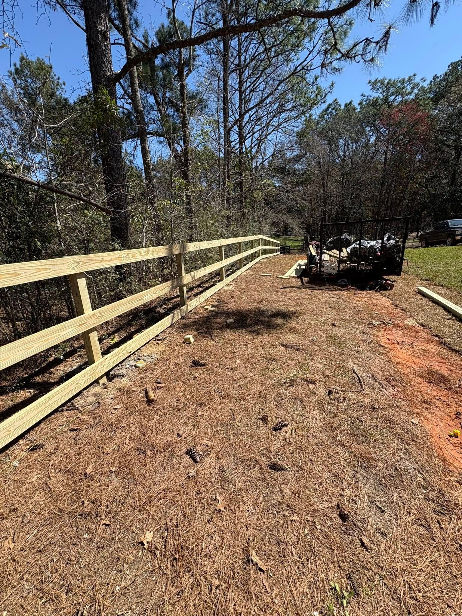 A newly installed wooden split-rail fence runs along a mulched path bordered by trees on a sunny day.
