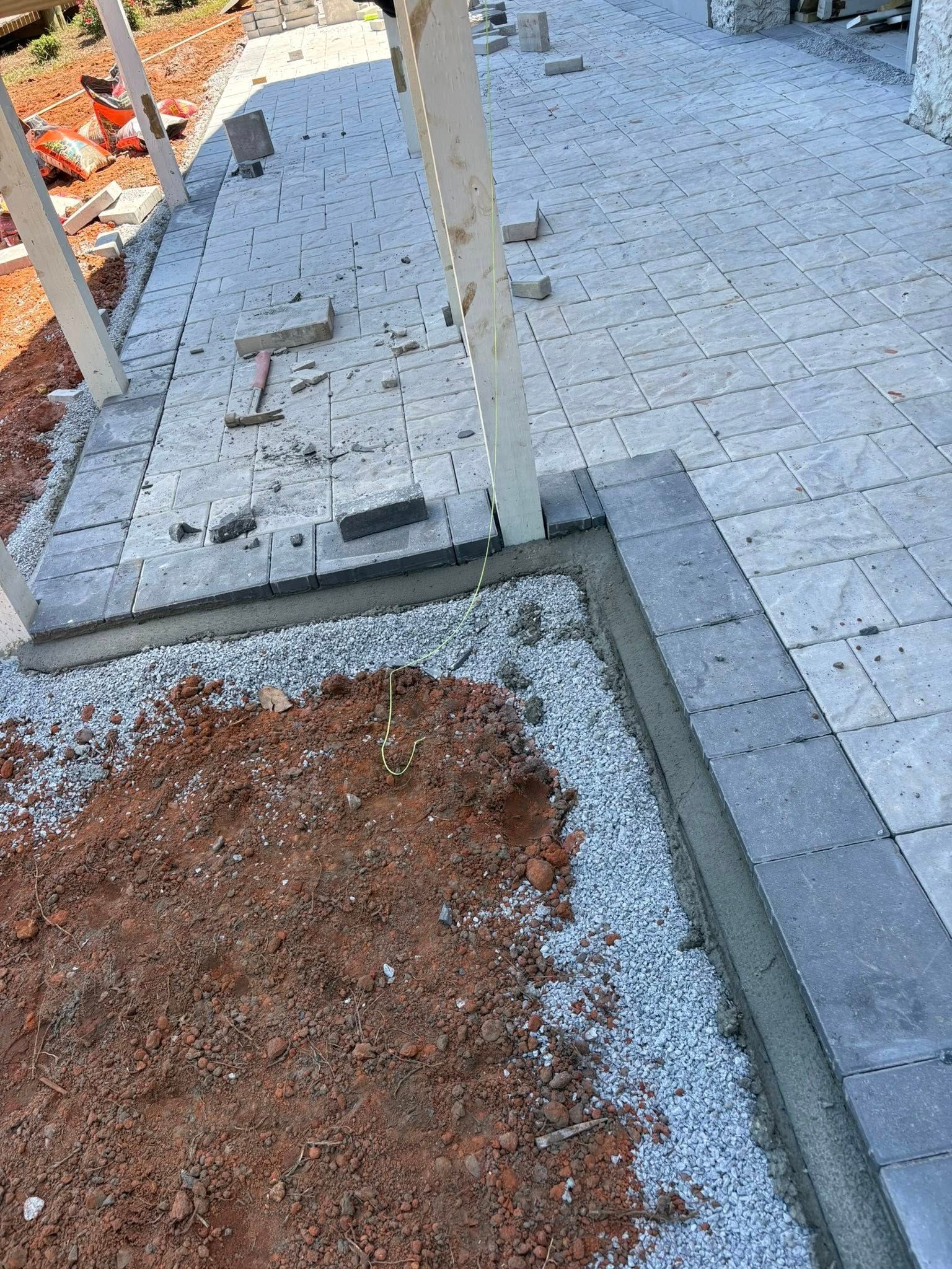 A high-angle view of a grey paver patio under construction, bordered by a mortar edge against loose gravel and red dirt.
