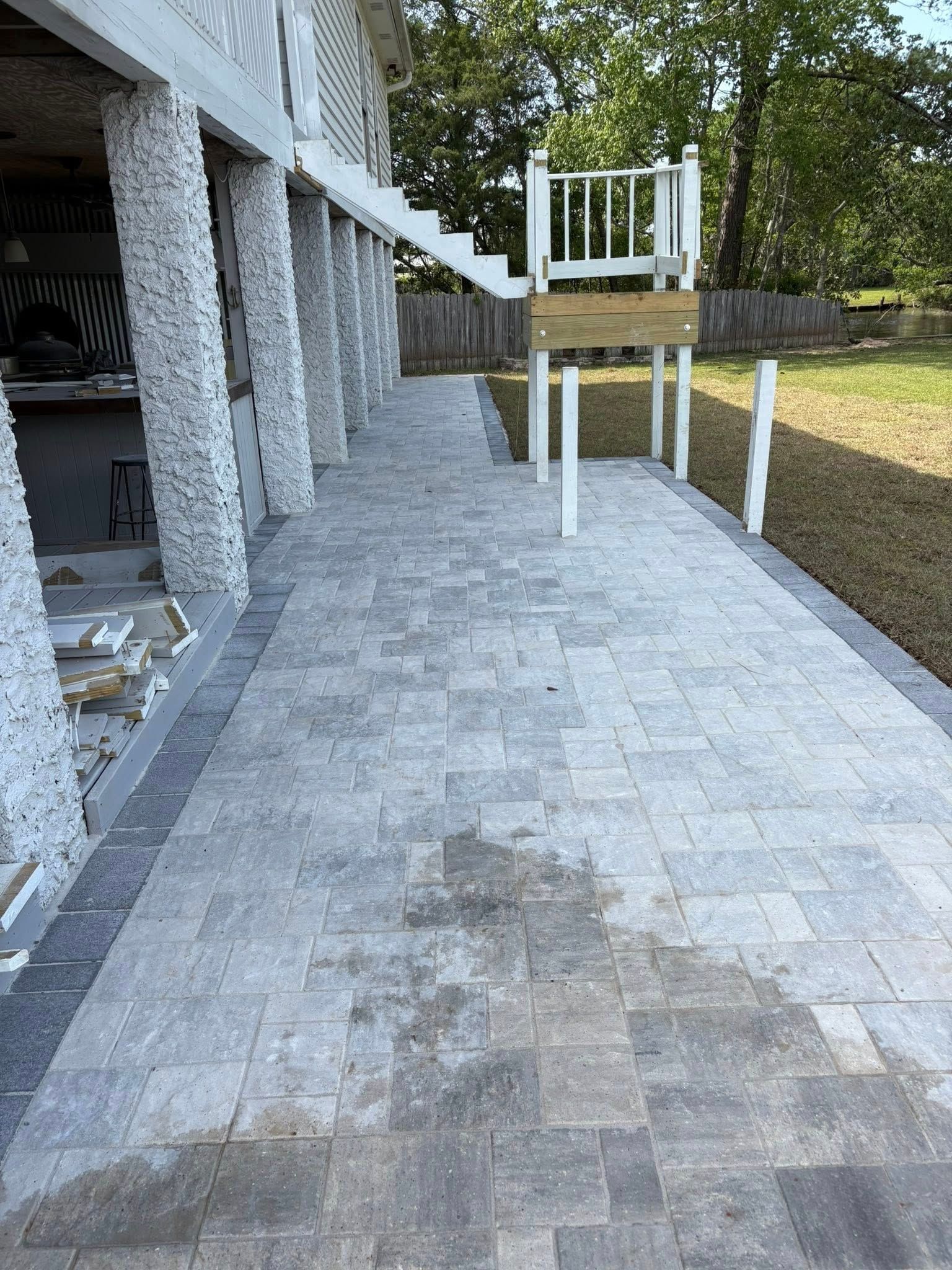 A paved patio area under a raised house structure with stone-textured columns, white stairs, and a wooden deck platform.