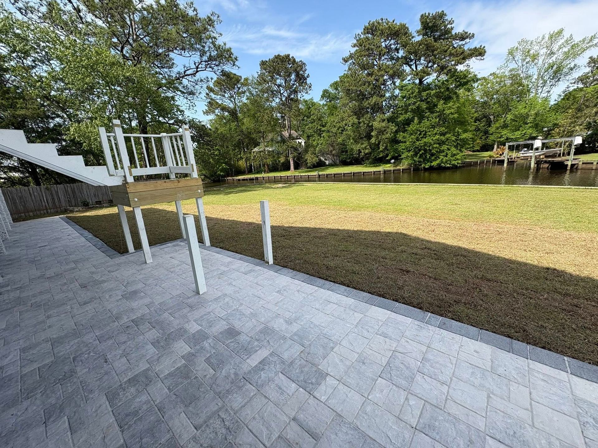 A gray paver patio overlooks a grassy backyard leading to a canal with a wooden dock and lush trees under a blue sky.