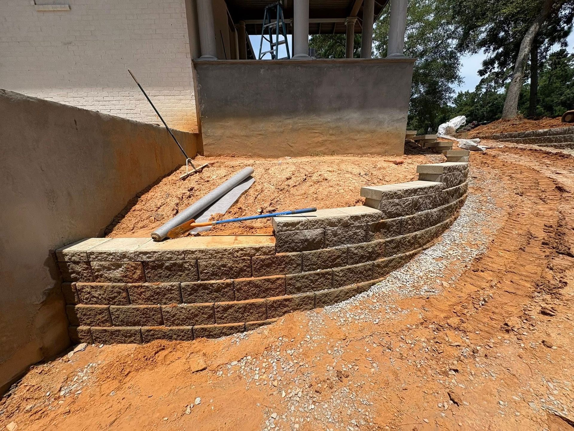 A curved, segmental retaining wall under construction in a yard with exposed soil, gravel, and building materials.