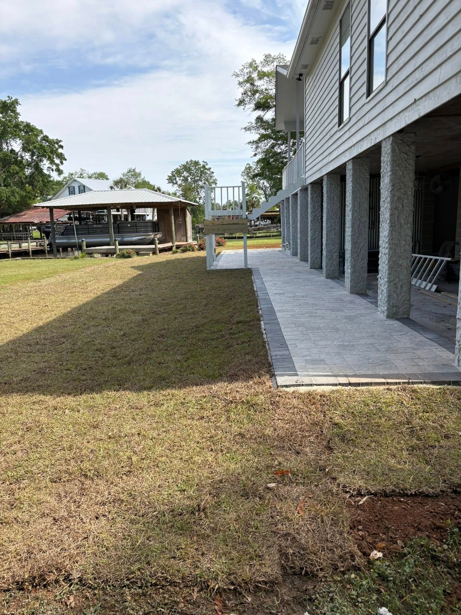 A view of a house raised on pillars with a new paved walkway beside a patch of recently laid sod in a grassy yard.