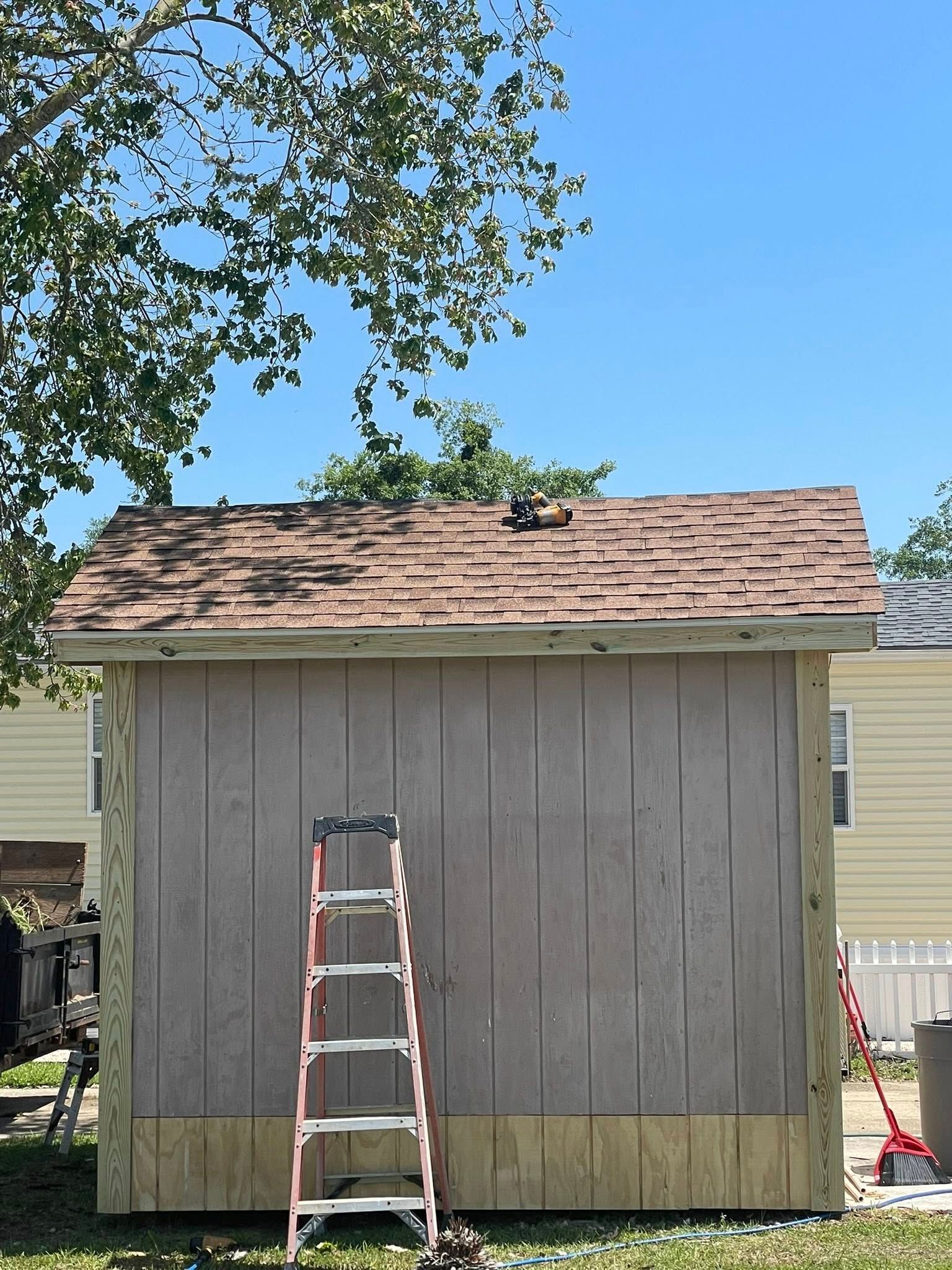 A wooden shed stands outdoors under a blue sky, featuring a newly shingled roof, a front-facing ladder, and side trim.