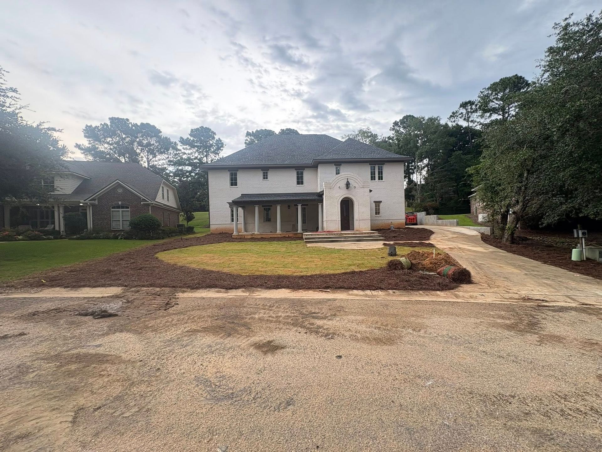 A white two-story house with a grey roof stands behind a freshly mulched yard and gravel driveway under a cloudy sky.