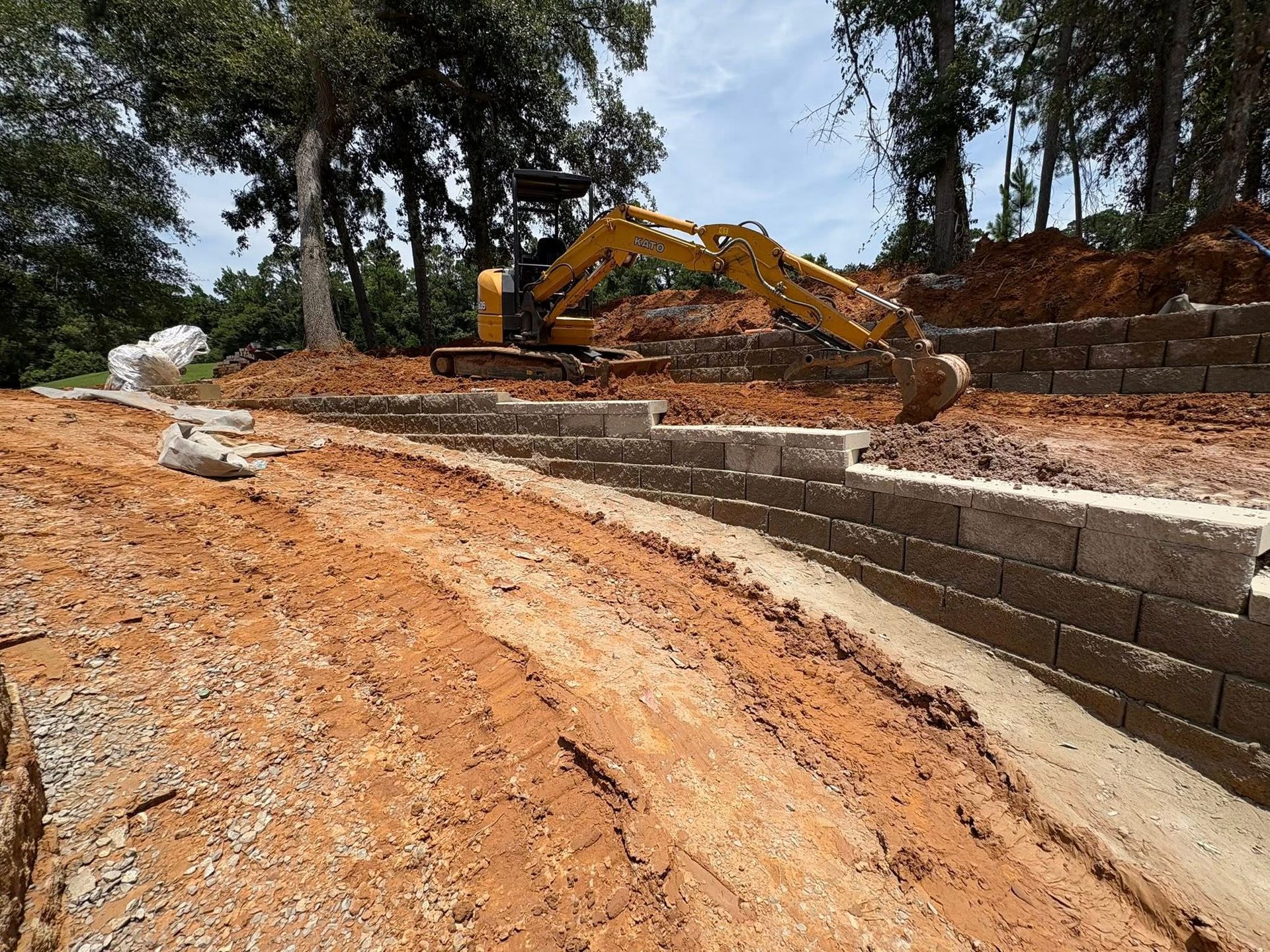 A yellow excavator sits on a dirt mound above a partially constructed stone retaining wall at a wooded construction site.