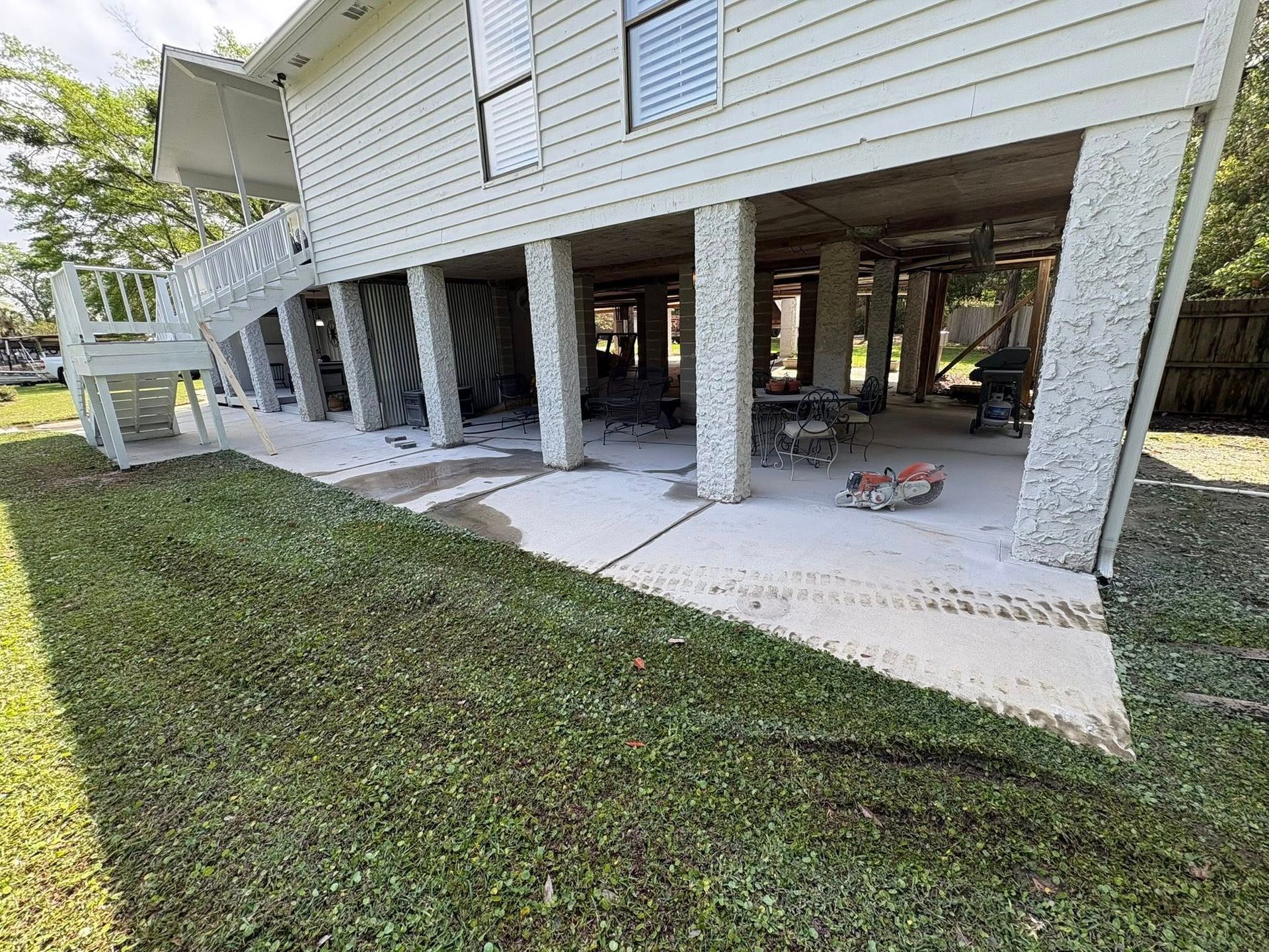 An elevated house with white siding stands on stone pillars above a concrete patio, with stairs leading to the first floor.