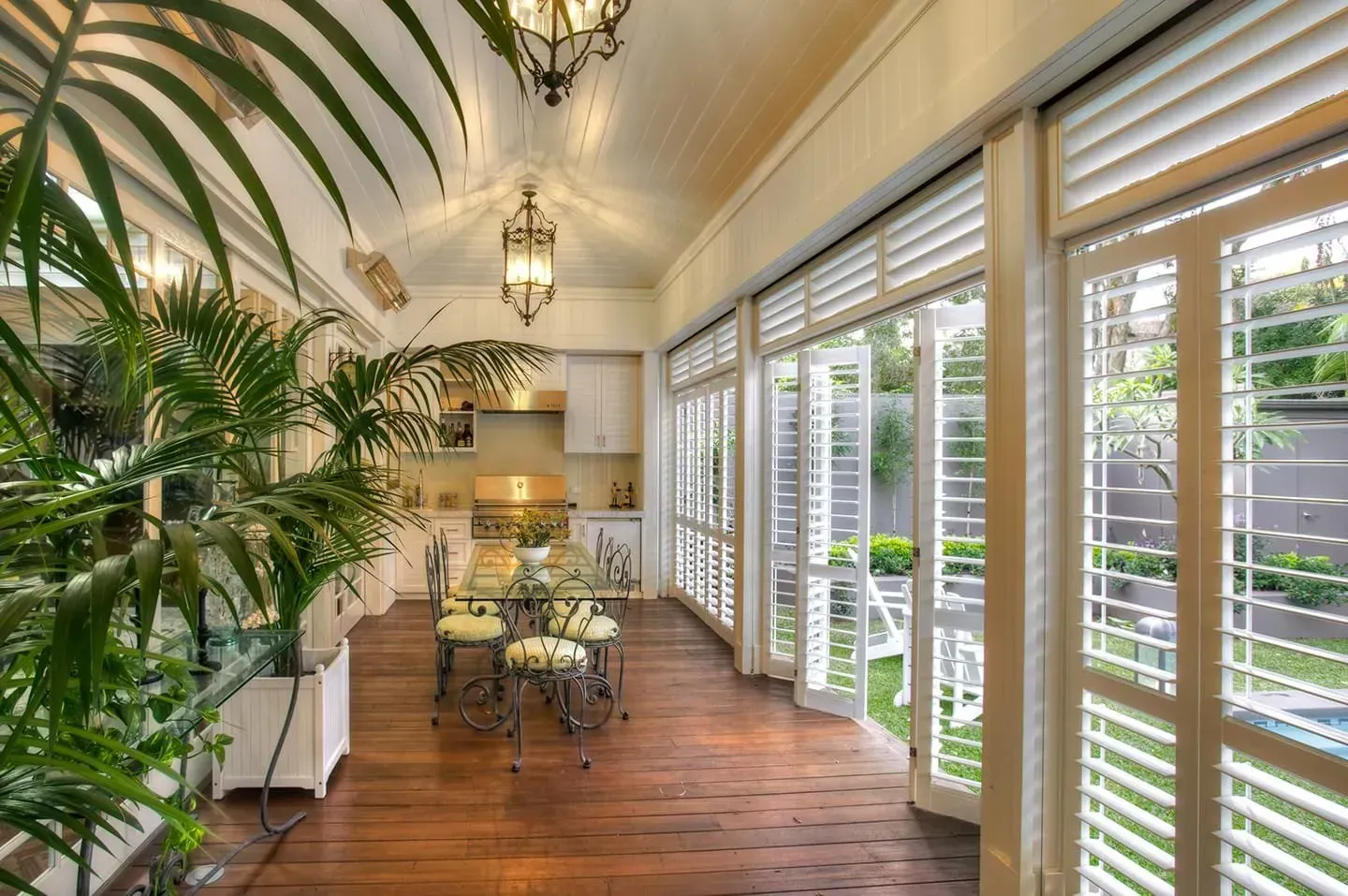 Sunroom with wooden floors, white shutters, and lush plants. A glass table and chairs are set inside.