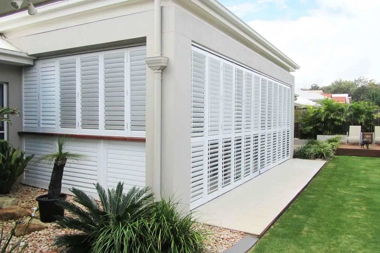 White shuttered exterior of a building with a lawn, plants, and concrete patio.