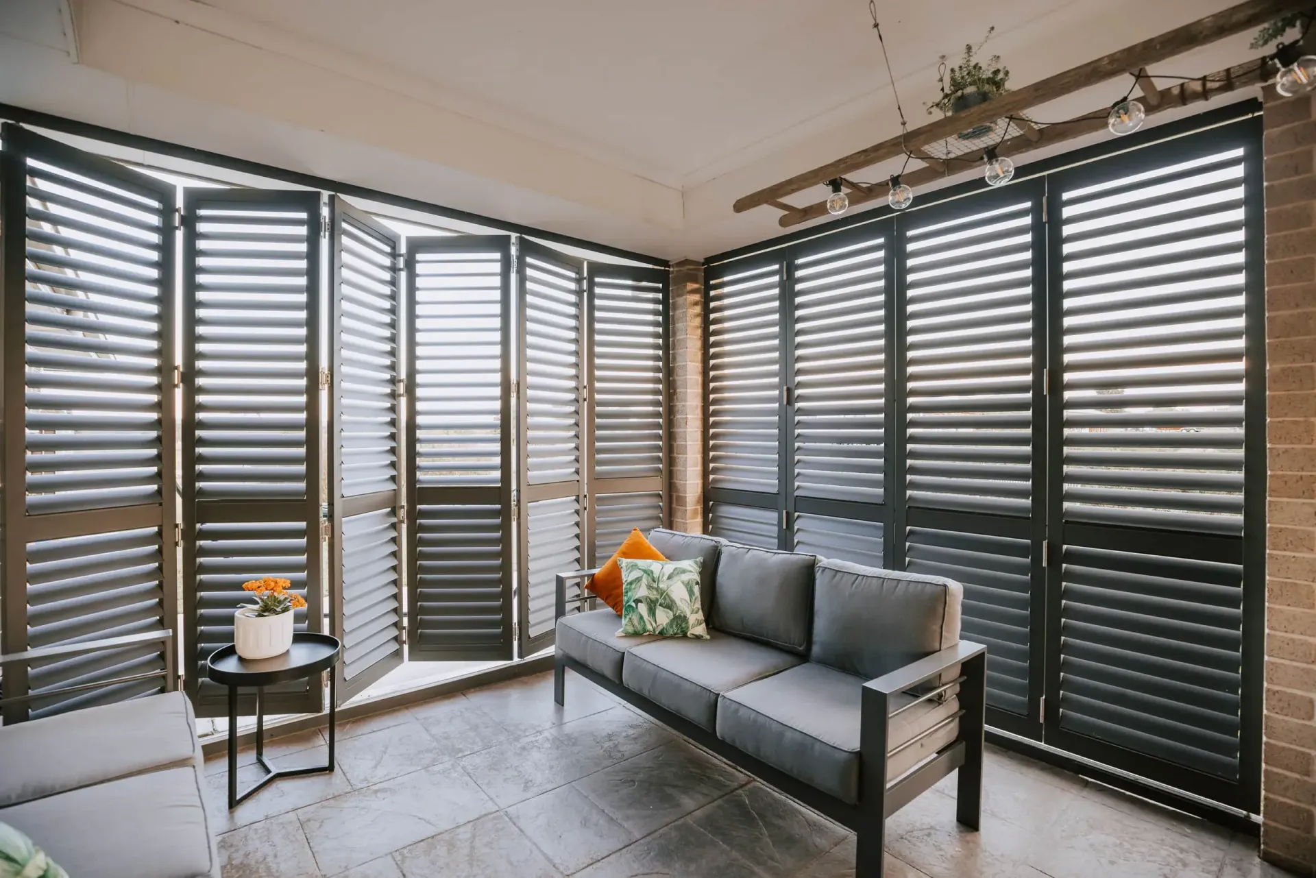 Sunroom with gray shutters, sofa, small table, and plants. Brick wall and bright natural light.