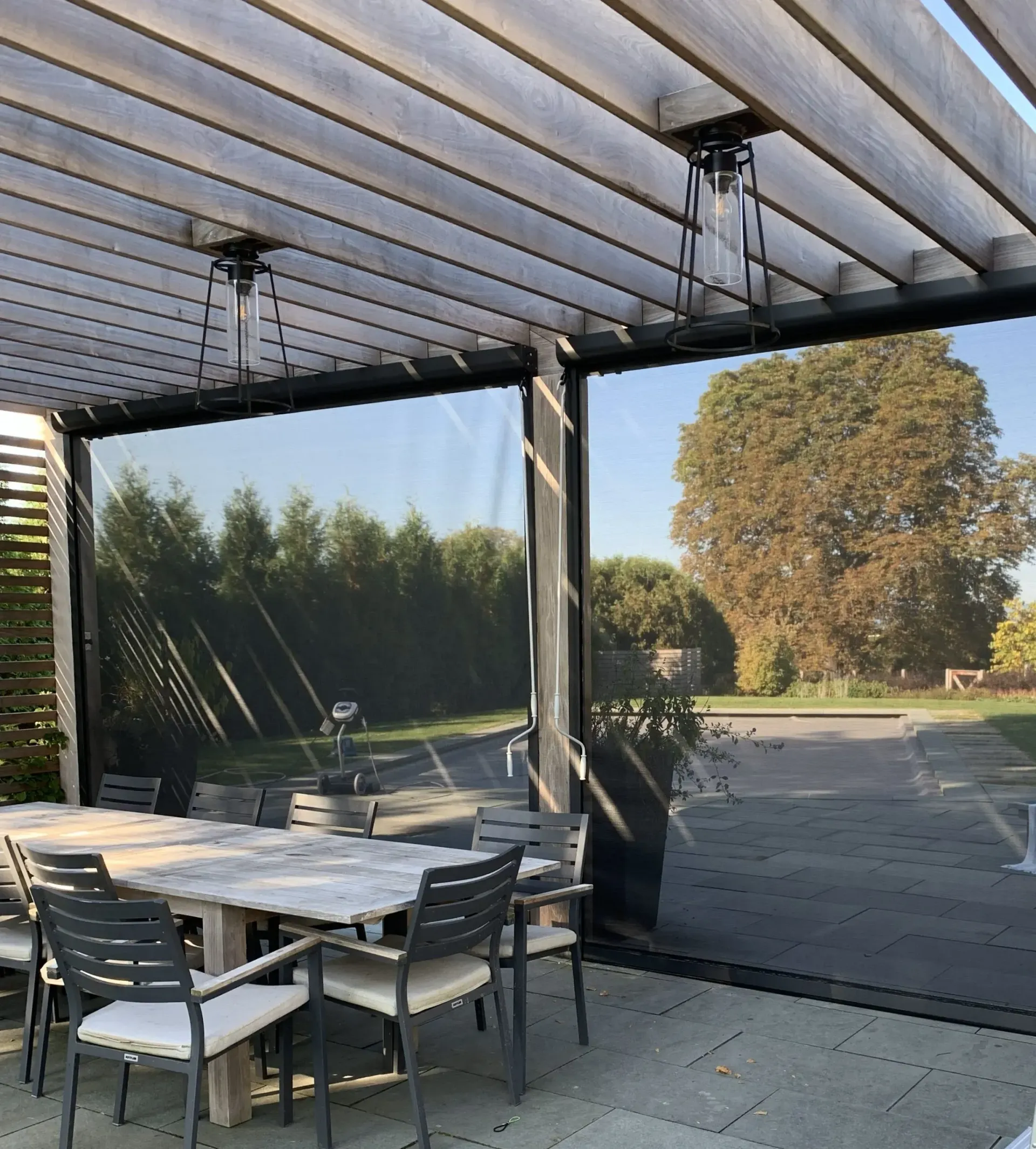 Patio with dining table, outdoor shades, and overhead pergola. Sunny day with trees in the background.