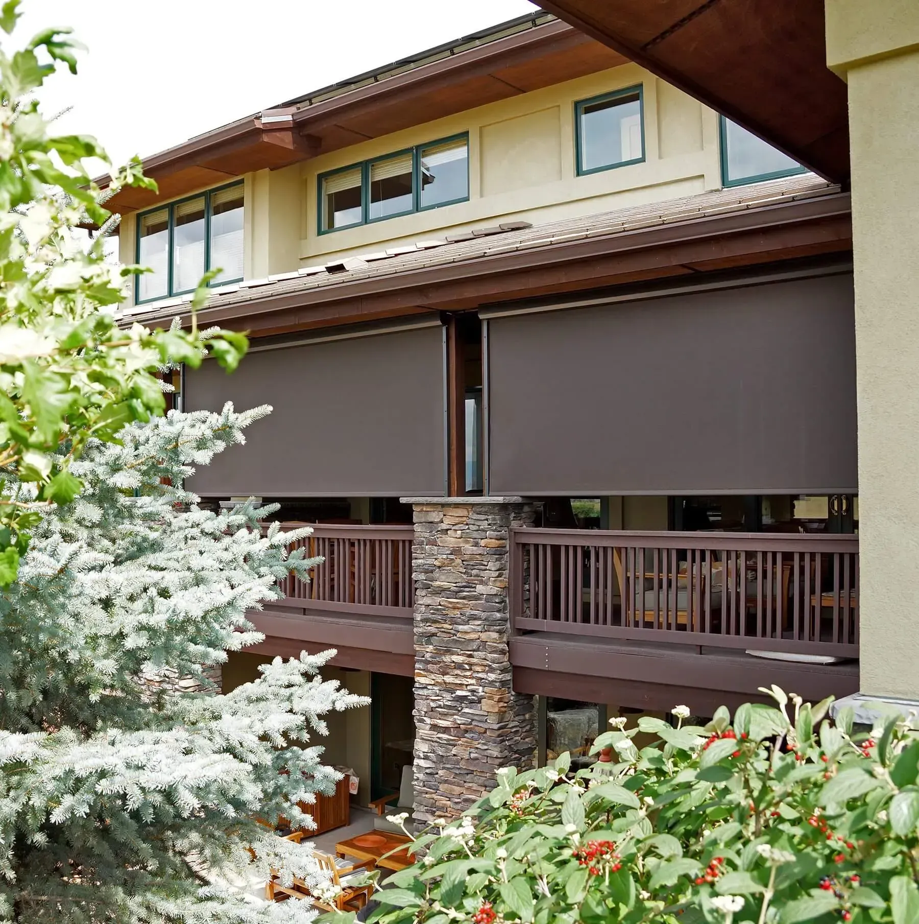 Brown outdoor shades on a house with a wooden deck, stone column, and greenery.