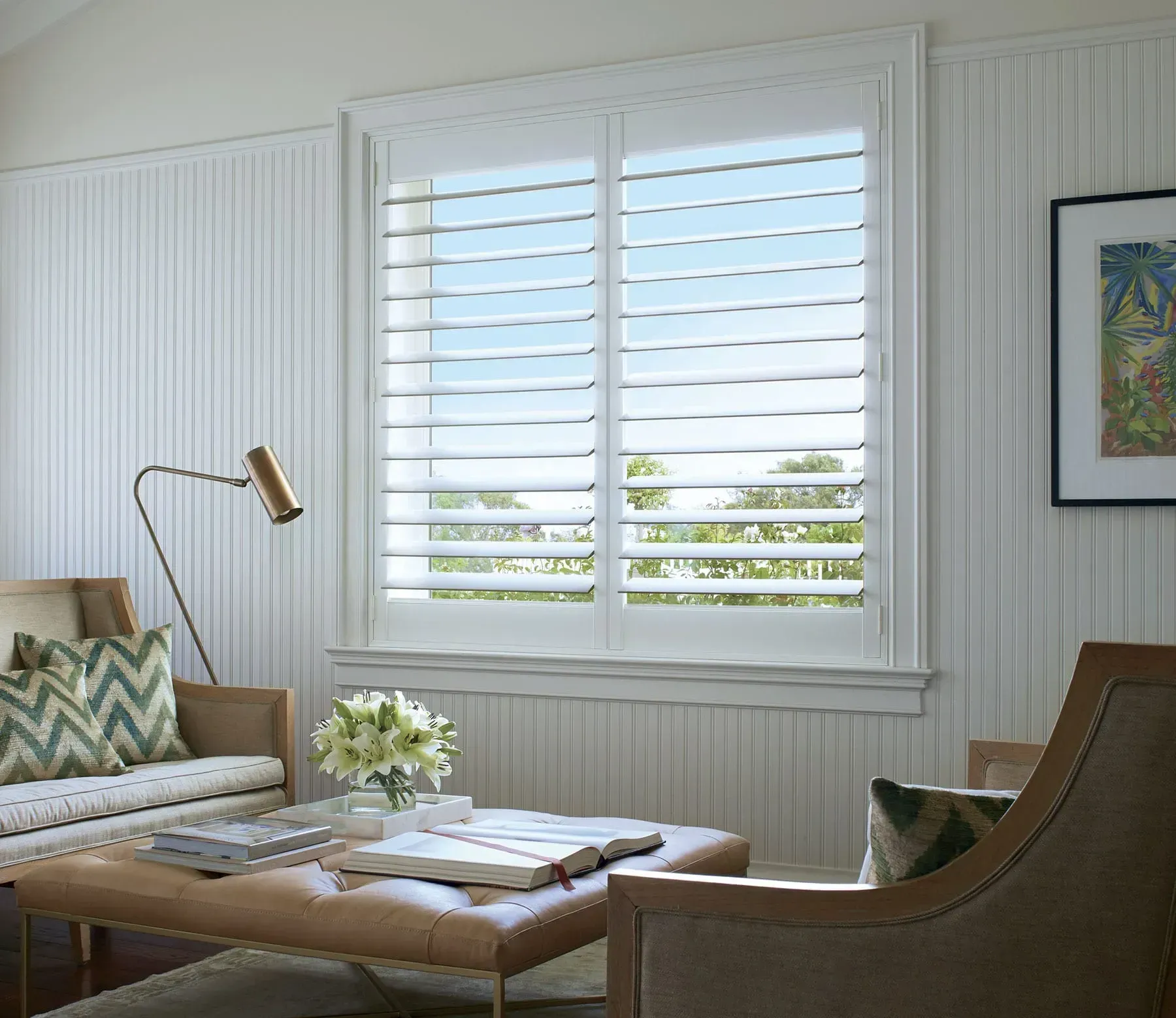 A bright room with white shutters, a couch, a chair, a lamp, and a framed picture on the wall.