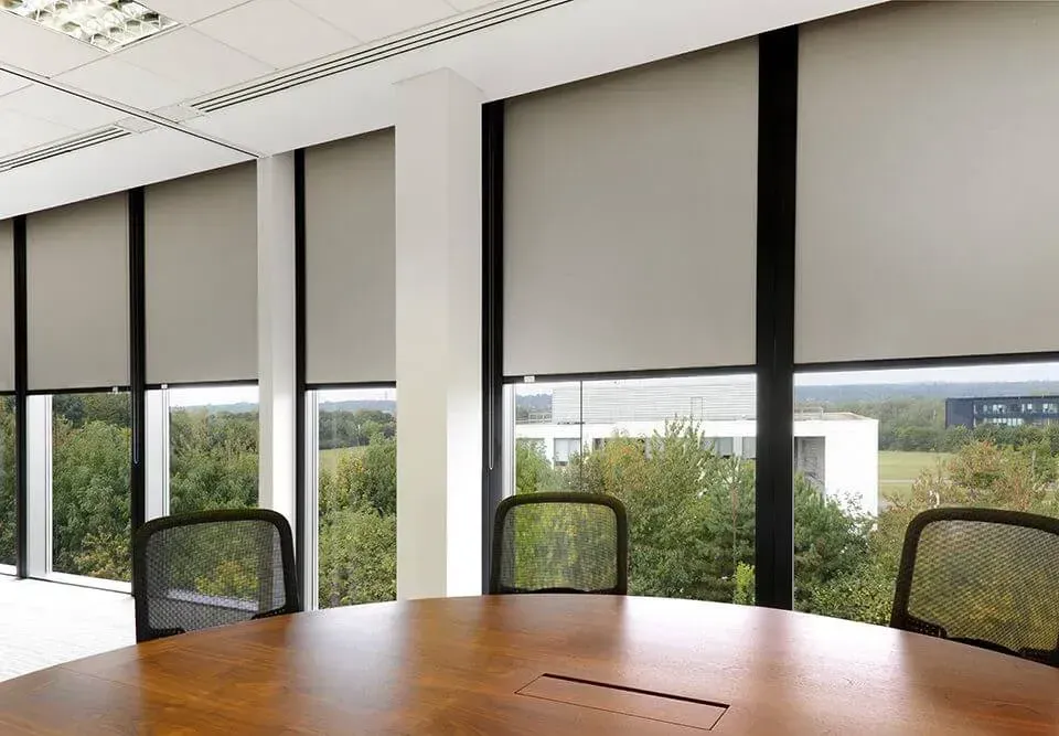 Conference room with a round table, chairs, and large windows with shades, overlooking trees.