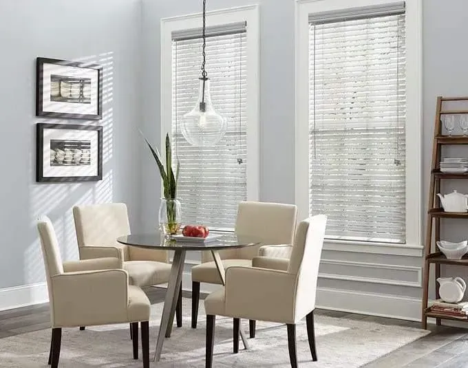Dining room with round table, four chairs, two windows with blinds, shelving unit, and artwork on wall.