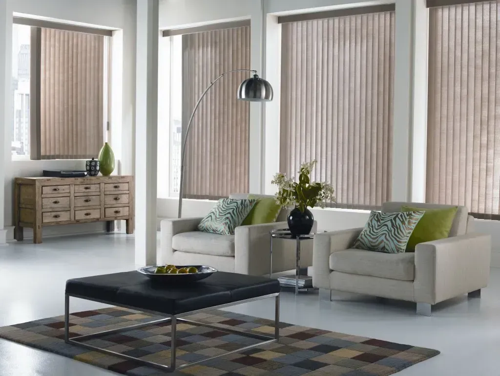Living room with beige vertical blinds, two cream armchairs, a rug, and a wooden chest.