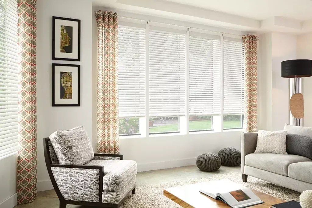 Living room with windows, blinds, patterned curtains, neutral furniture, and art.