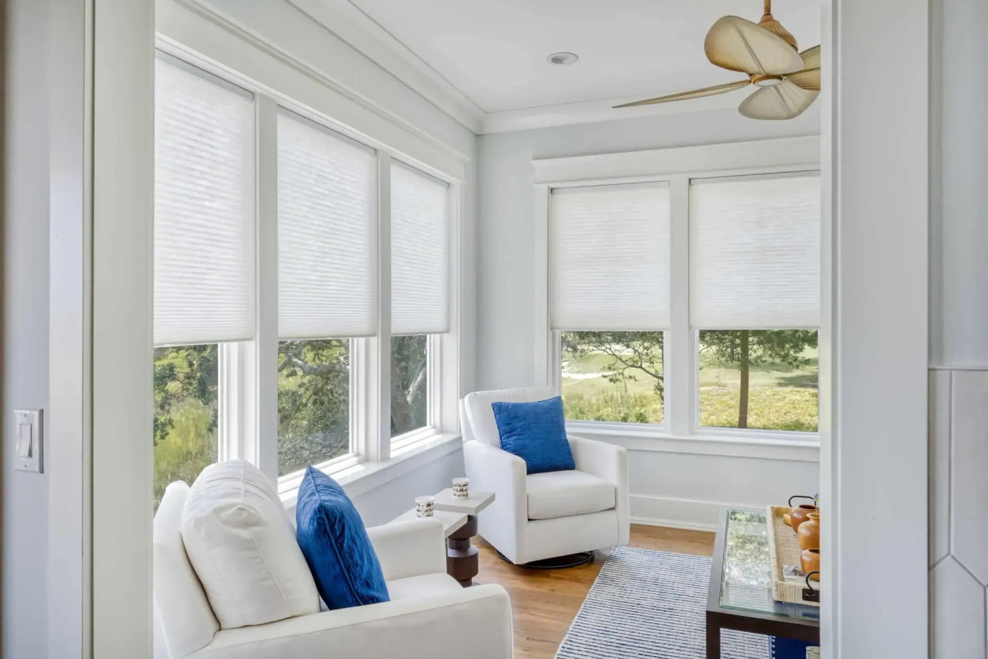 Cozy sunroom with white chairs, blue pillows, and windows with white blinds, overlooking a green landscape.