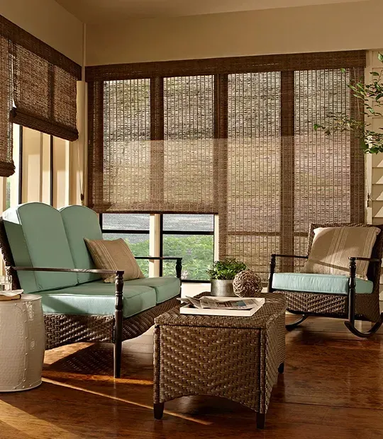 Sunroom with wicker furniture, light blue cushions, and woven shades. Wooden floors.