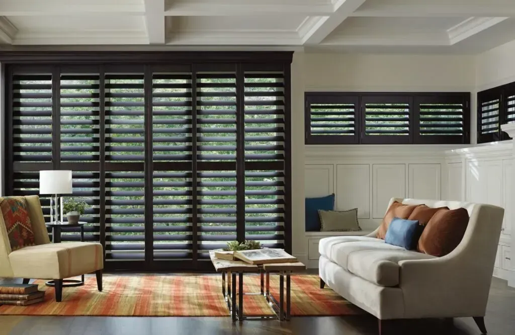 Living room with dark wood shutters, beige sofa, colorful rug, and white walls.
