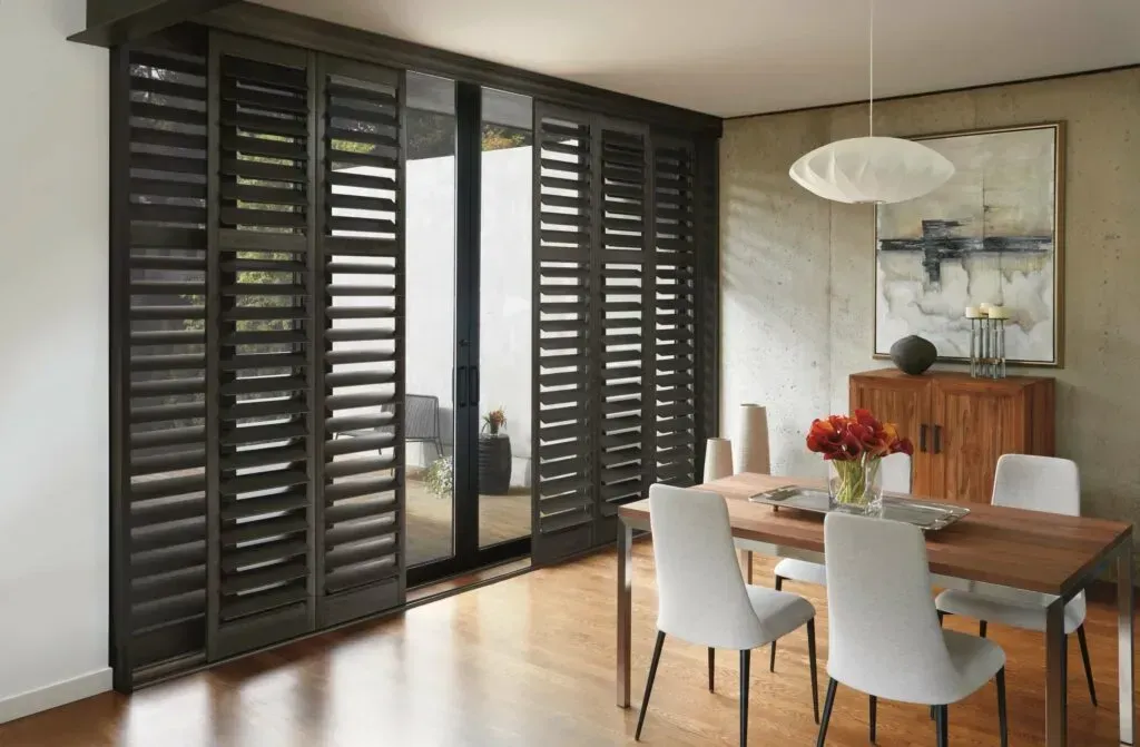Dark wooden shutters covering a sliding glass door, dining room with table, chairs, art, and light fixture.