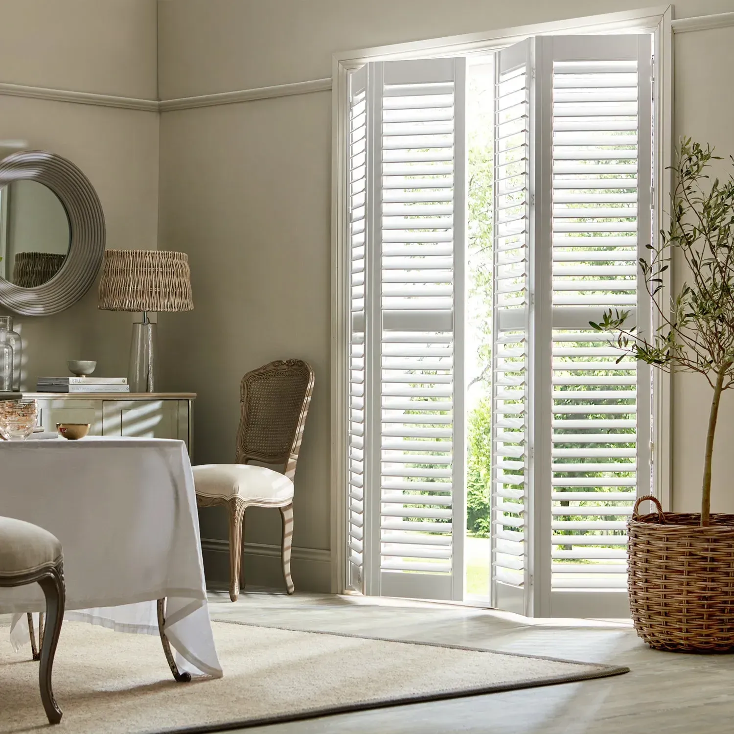 Dining room with white shutters, table, chairs, mirror, and potted tree. Natural light streams in.