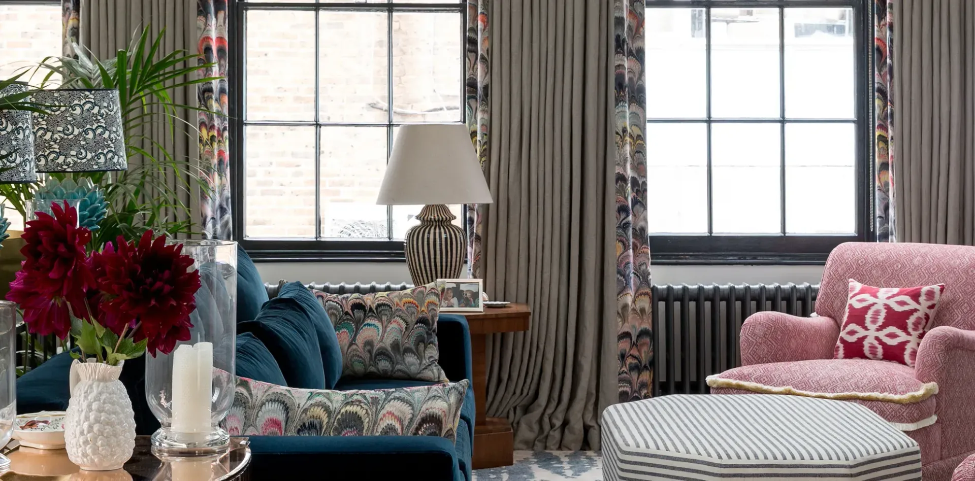 Cozy living room with floral curtains, blue sofa, pink armchair, and striped ottoman.