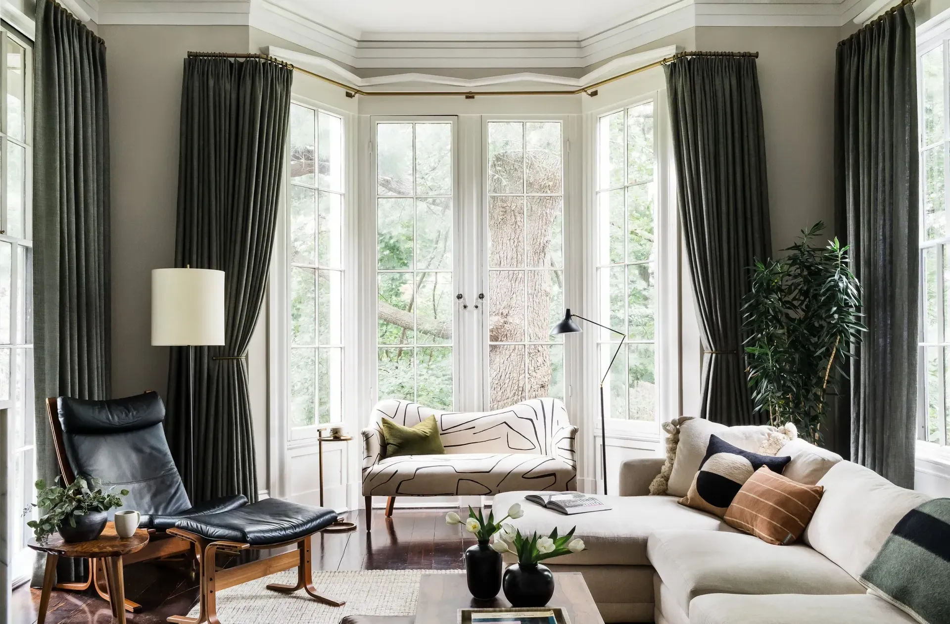 Living room with bay window, ivory sectional, black leather chair, dark green curtains, and natural light.