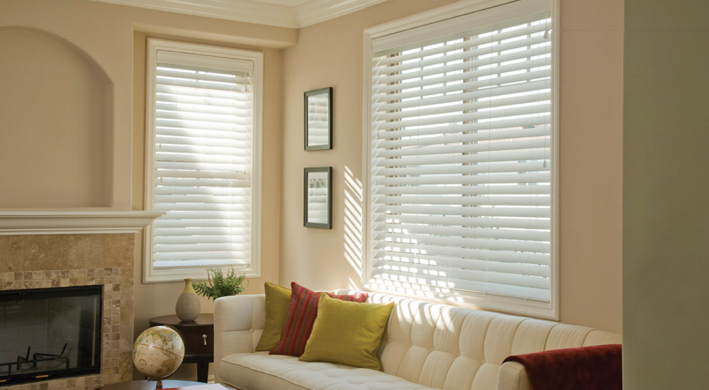 Living room with white sofa, fireplace, and two windows with blinds, sunlight streaming in.