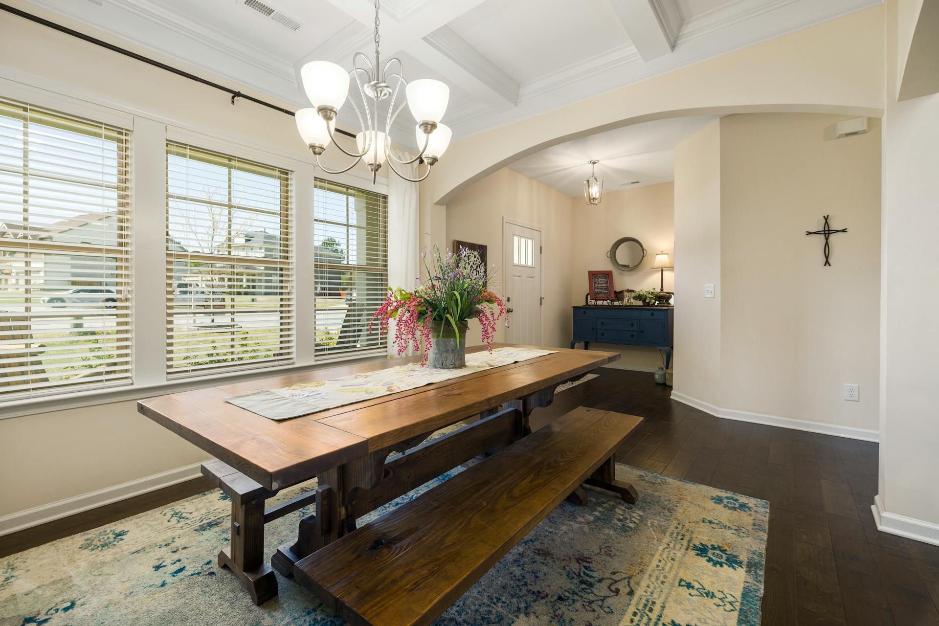 Dining room with wooden table, bench, chandelier, windows, rug, and an arched doorway.