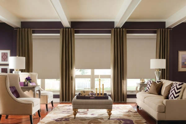 Living room with beige furniture, patterned rug, and brown curtains framing three windows with tan blinds.