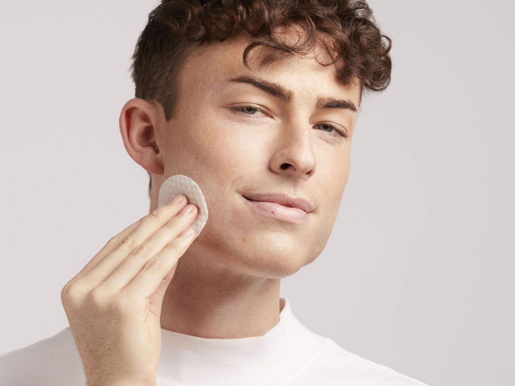 A man with curly hair uses a cotton pad on his cheek.