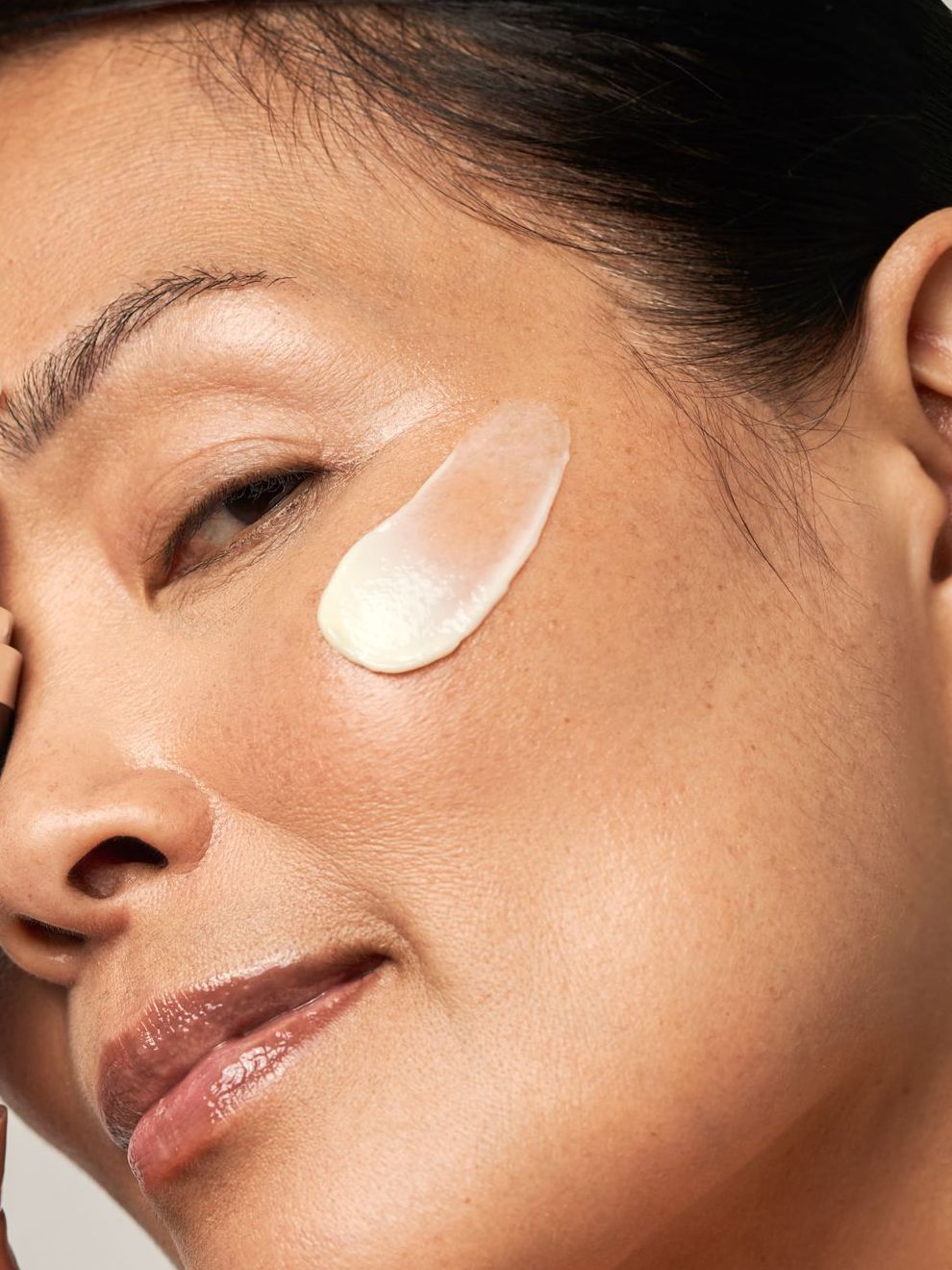 Woman's face with cream applied to cheek. She smiles, showing skin texture and a light-colored product.