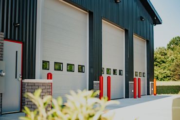 Industrial building with three white roll-up doors, red bollards, and a sunlit concrete driveway
