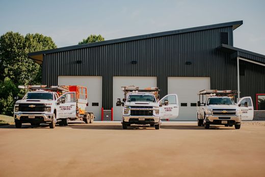 Three utility trucks parked outside a dark metal garage with open bays