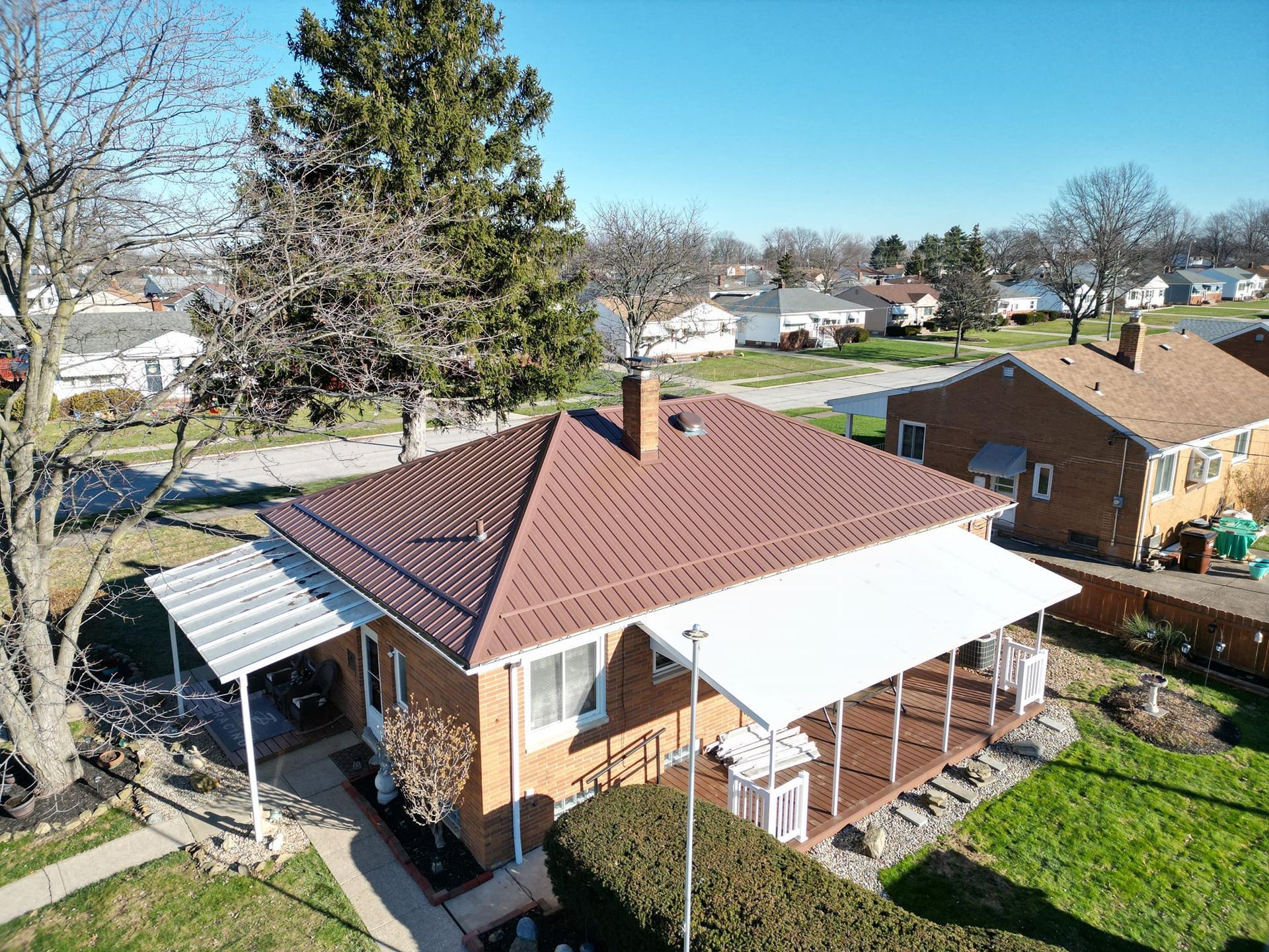 Aerial view of a brick house with a brown roof and awnings, in a suburban neighborhood on a sunny day.
