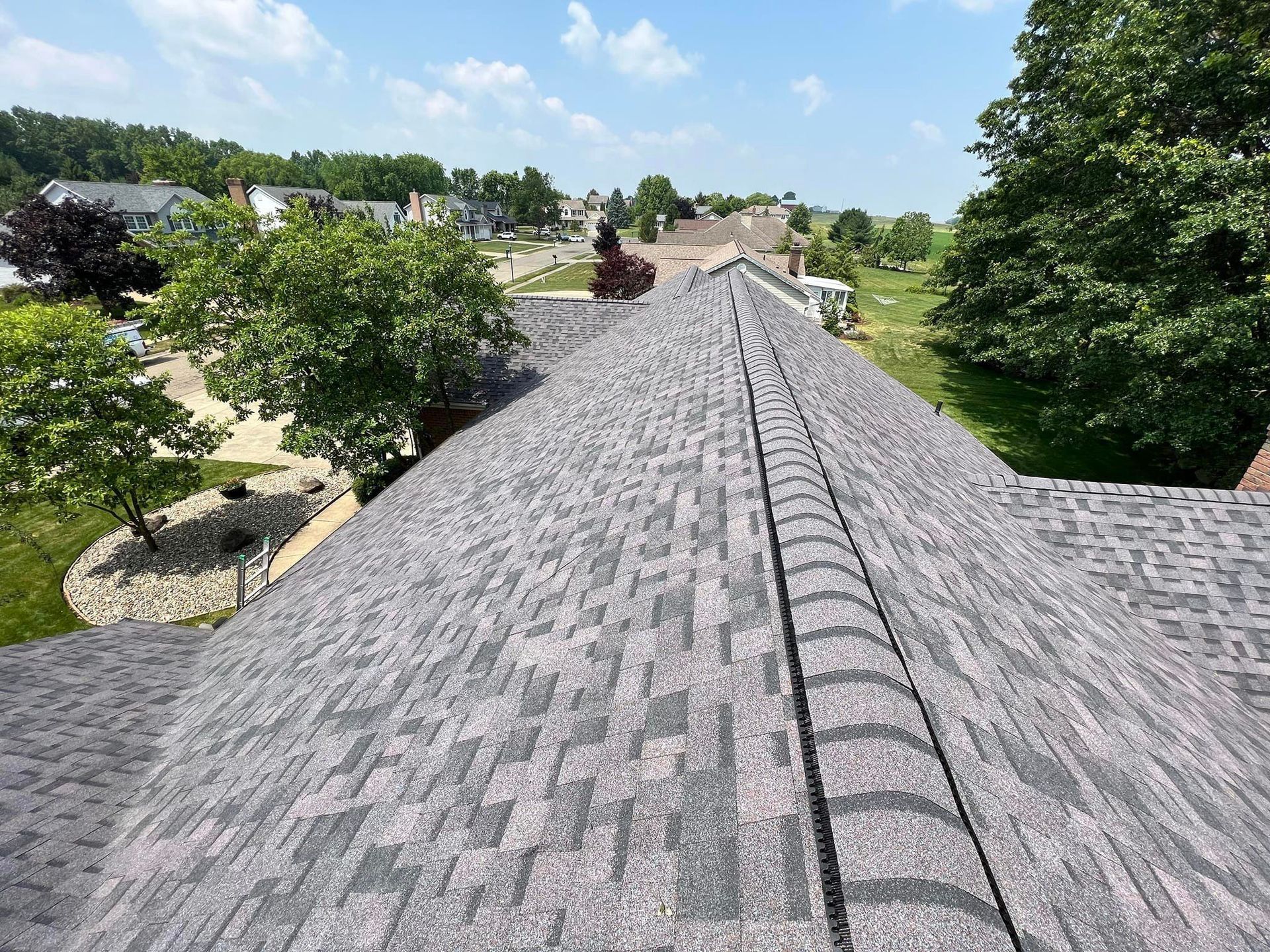 View from a roof of a residential area with a gray shingled roof and trees. Blue sky visible.