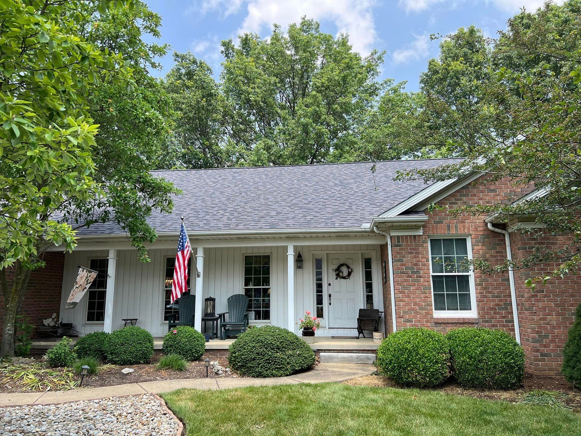 Brick house with a white porch, front door, and American flag; green bushes and trees surround it.