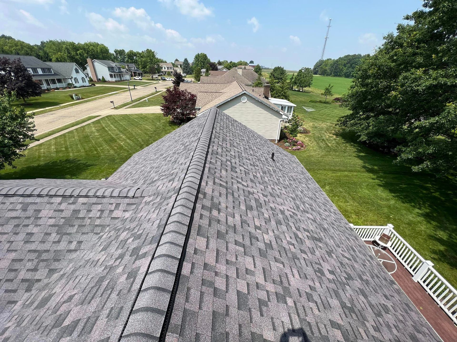 Overhead view of a gray shingled roof with a green lawn, houses, and trees visible in the background under a blue sky.