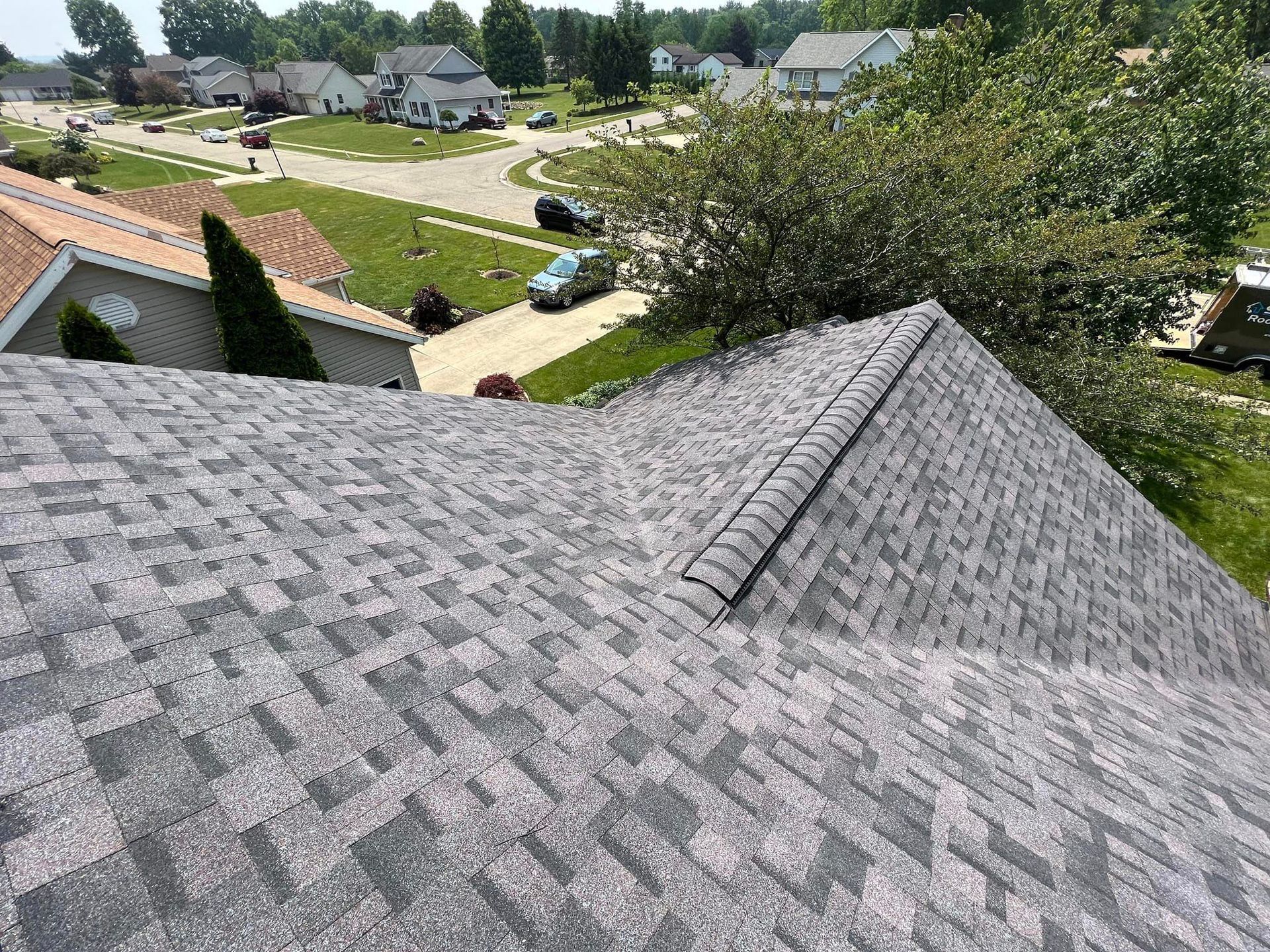 View of a gray asphalt shingle roof with a neighborhood in the background on a sunny day.