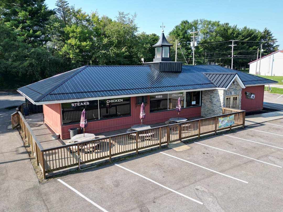 A red brick building with a black roof and a deck in front of a parking lot, with a small steeple.