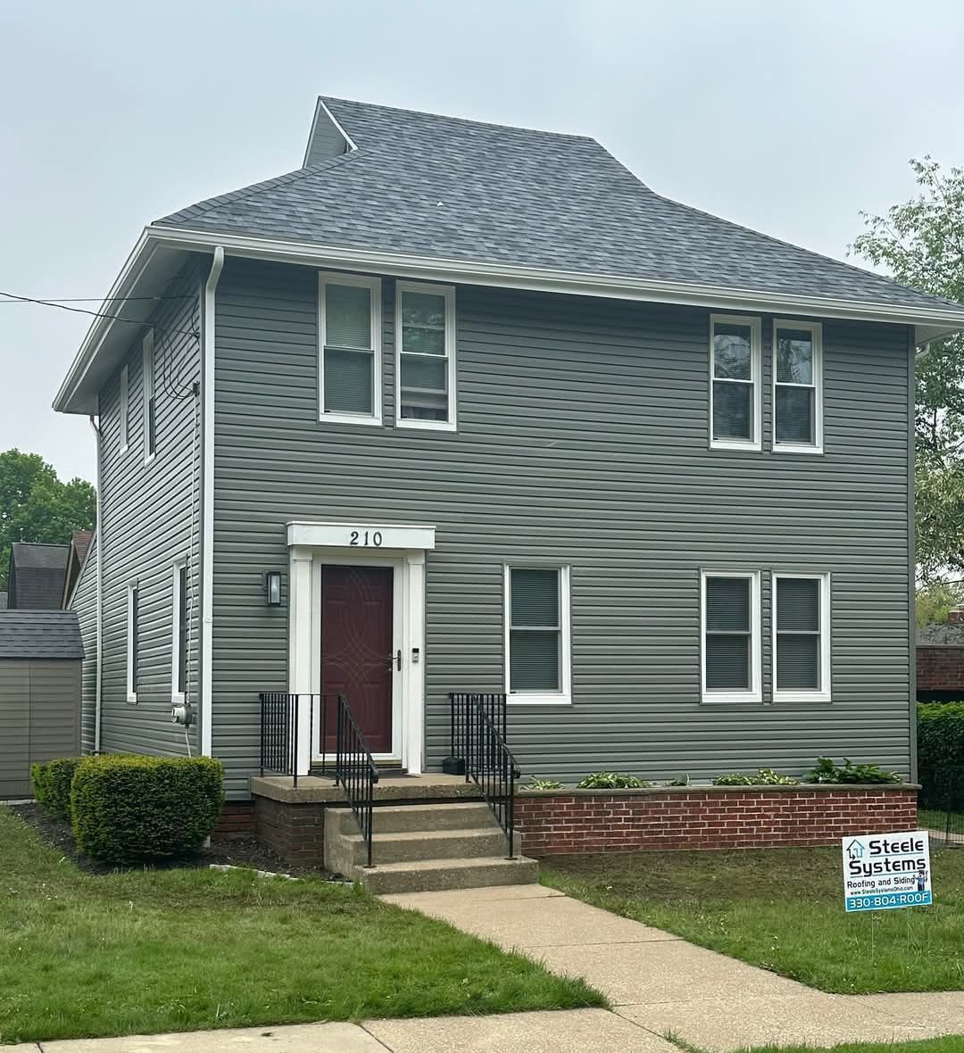 Two-story house with gray siding, dark red door, and a brick foundation. 