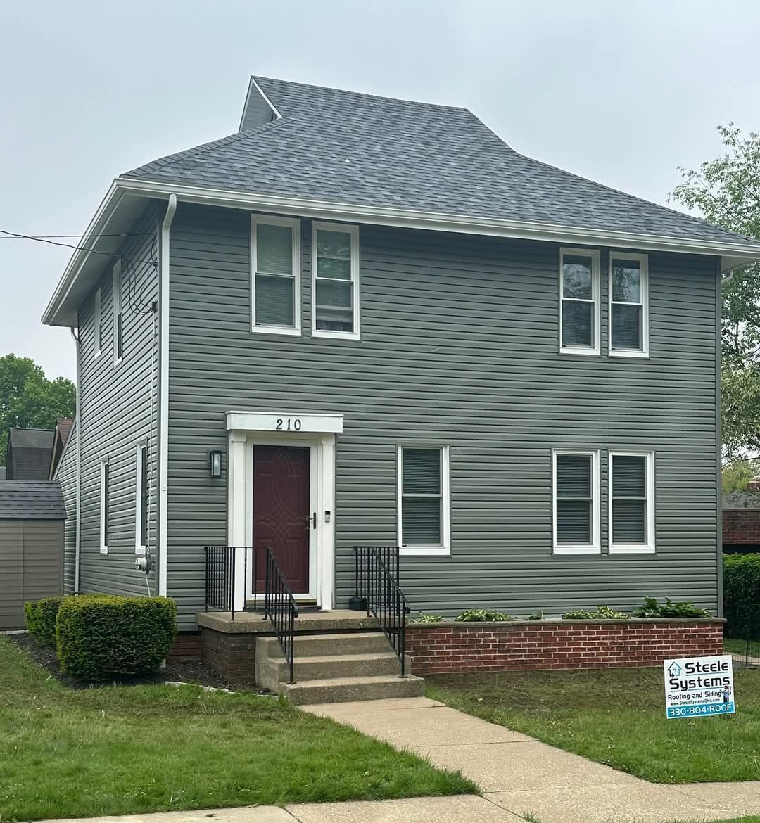 Two-story house with gray siding and a unique roof, featuring a red door and a small front lawn with a sign.