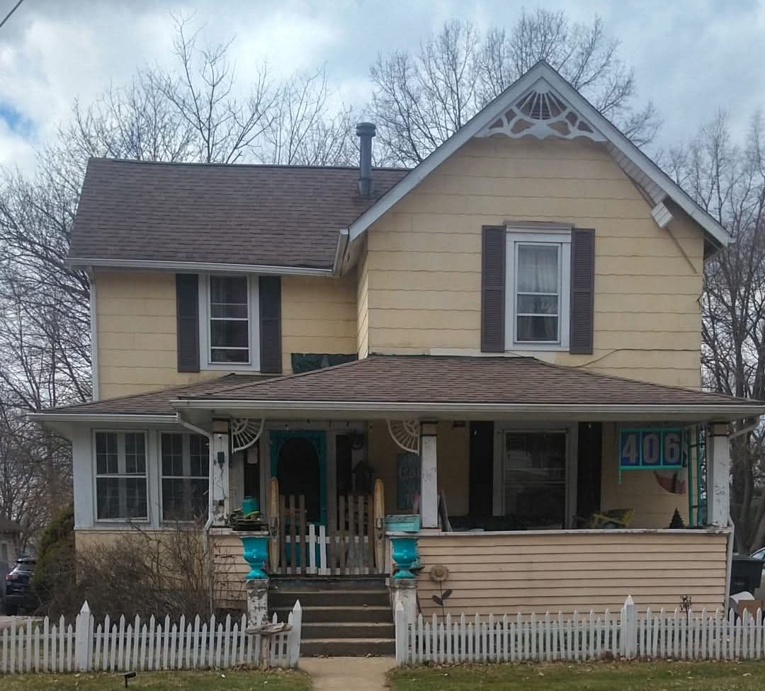 Two-story yellow house with a porch, white picket fence, and brown roof. 