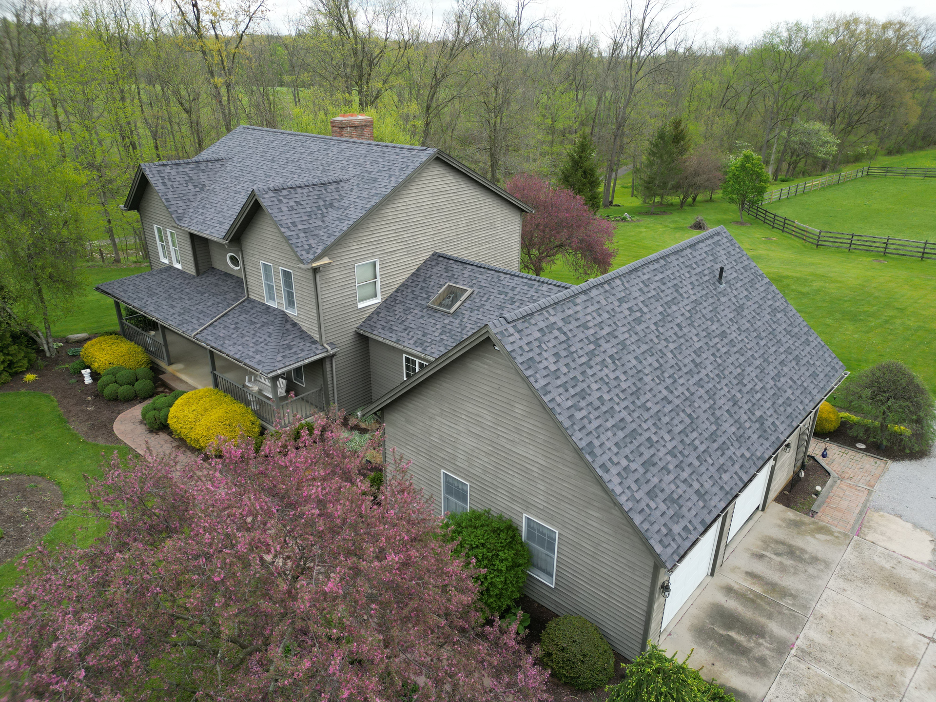 An aerial view of a gray house with a dark gray roof and attached garage, surrounded by trees and green grass.