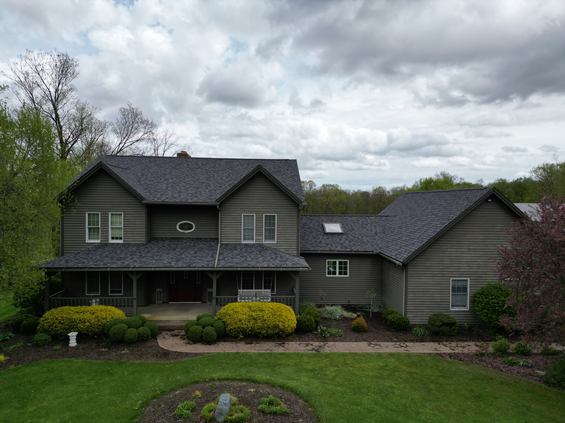 A gray two-story house with a wraparound porch, surrounded by greenery under a cloudy sky.