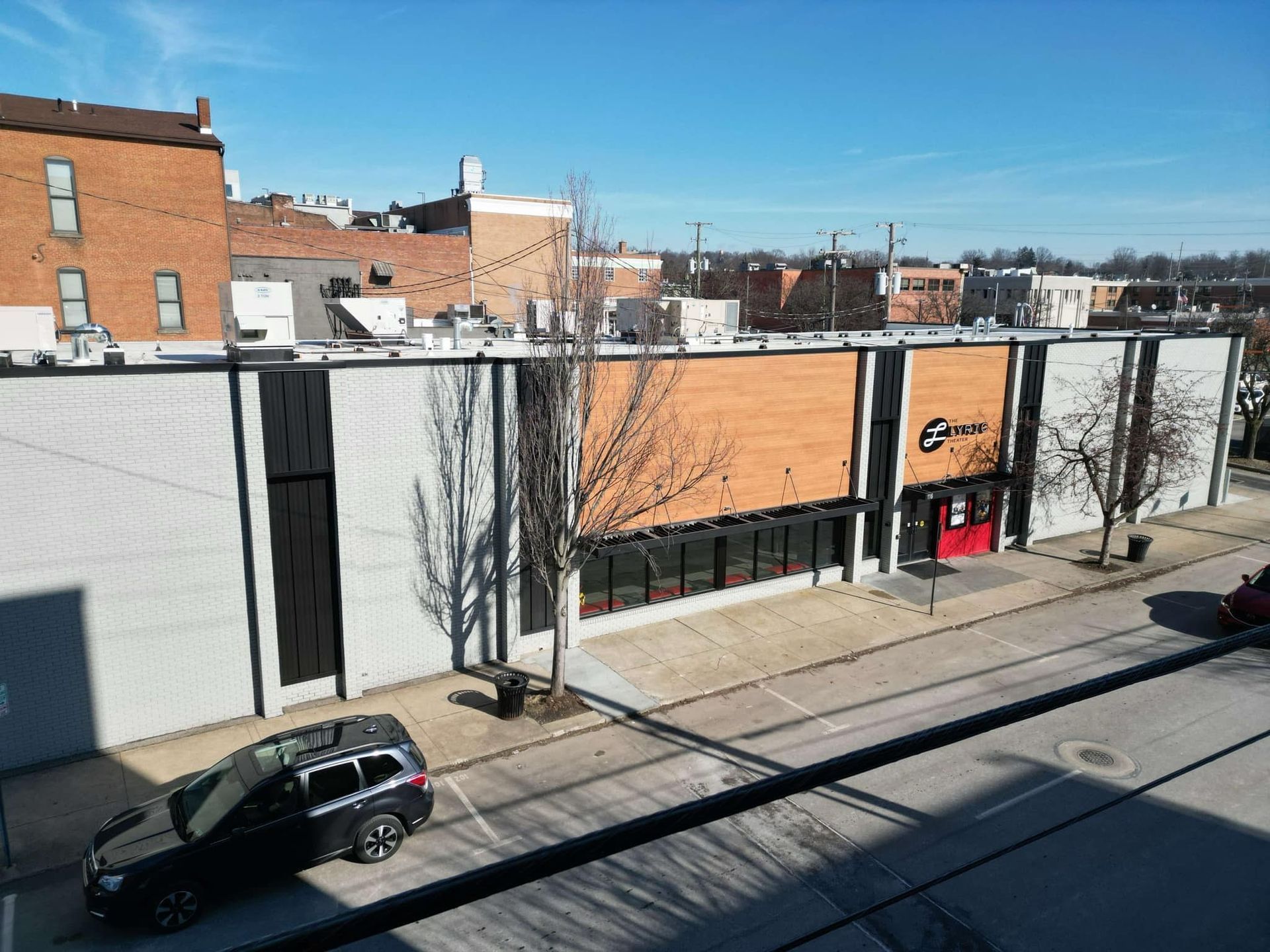 Exterior view of a commercial building with a brick facade and a red entrance. A black SUV is parked on the street in front.